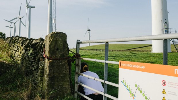 Composer Sara Rahman crouches just inside the gate of a wind farm in Yorkshire. The wind turbines are visible in the image and span a lush green pasture bordered by a jagged stone wall.