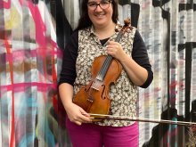 Composer Rachael Hales standing in front of a wall of graffiti holding a violin