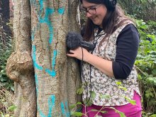 Composer Rachael Hales standing close to a large tree trunk, with forehead and handheld sound recorder in contact with the tree