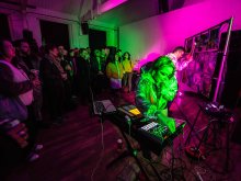 This photo shows Lia Mice performing live onstage playing an Ableton Push and singing into a microphone. Behind her, Andrew Booker plays Lia's large instrument The Catastrophone.